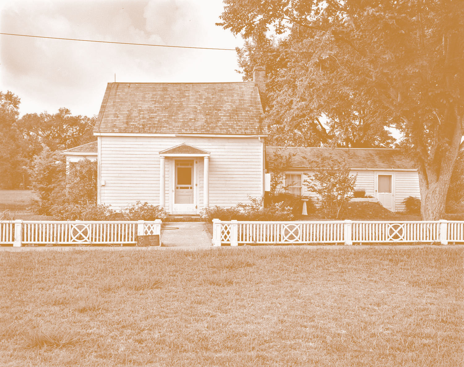 Sepia-toned house with white siding and gray shingled roof, wooden fence, trees, and grass