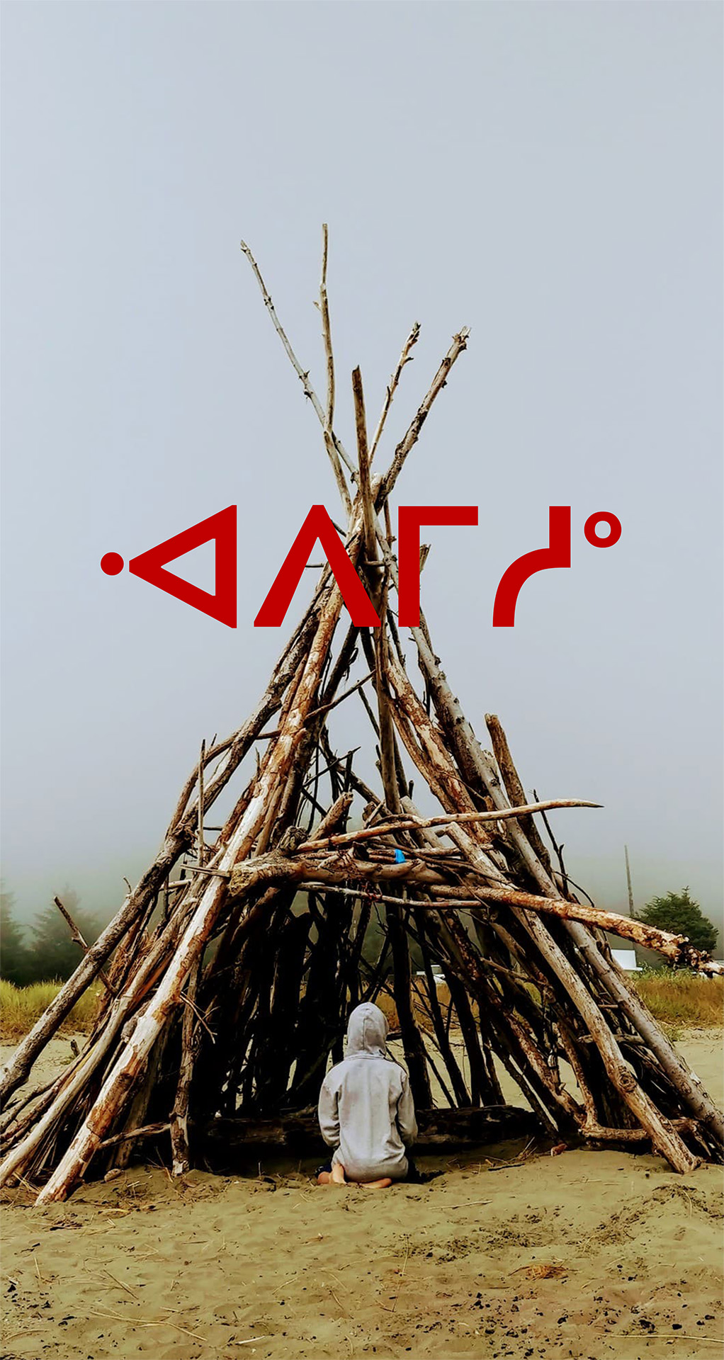 A person with their back to the camera sits in a wooden tipi on sand. Cree syllabics are overlayed